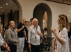 A group of adults, some wearing lanyards, listen to a woman speaking in an indoor space with arches and a stroller in the background.
