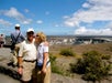 A woman kisses a man on the cheek while posing for a photo at a scenic overlook with a crater and blue sky in the background. Other people are also sightseeing.