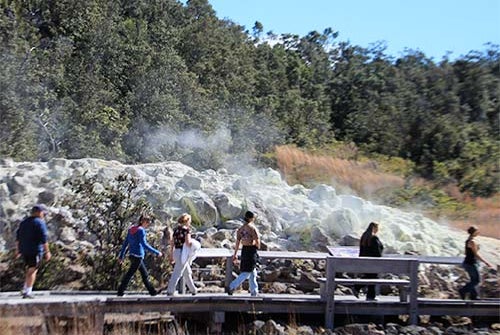 A group of people walk on a wooden boardwalk past steaming rocks in a natural outdoor setting with trees in the background.