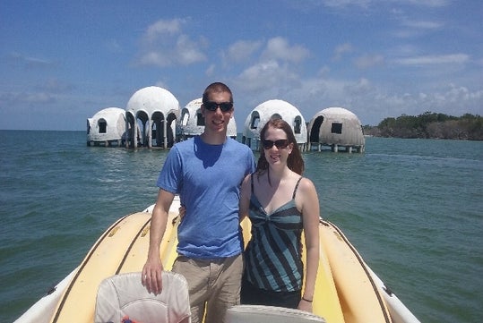 A man and woman stand on a yellow boat in front of dome-shaped houses on the water under a partly cloudy sky.