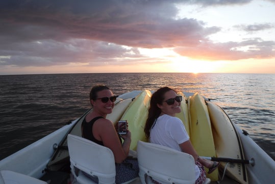 Two people sit on a boat with kayaks at sunset, smiling toward the camera while the sun sets over the ocean in the background.