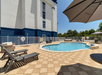 Outdoor swimming pool with lounge chairs and large umbrella on a tiled patio, enclosed by a white fence. Building and greenery in the background under a clear blue sky.