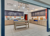 Breakfast buffet area with wooden cabinets, beverage dispensers, and a center island displaying food. Large chalkboard-style decor on walls. Modern lighting fixtures hanging from the ceiling.