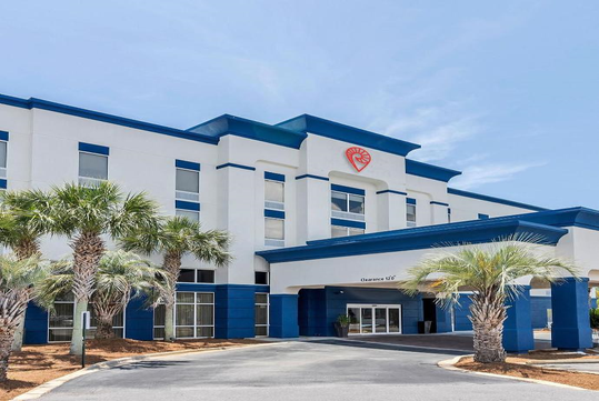 Exterior of a white and blue hotel building with a red heart logo, surrounded by palm trees, under a clear blue sky.