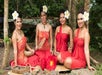 Four women in red dresses with flower hair accessories sit on a mat outdoors, holding woven fans.