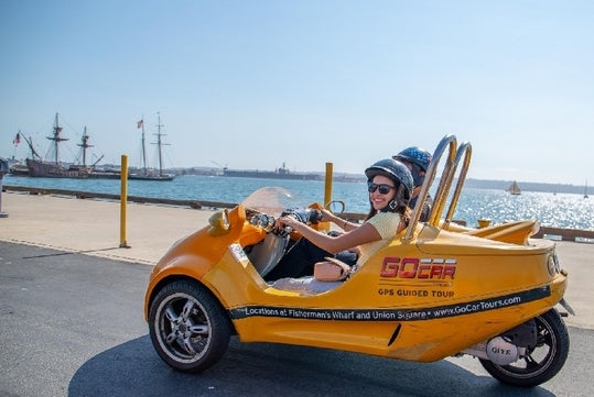 Two people ride a yellow GoCar near a waterfront with a sailing ship in the background.