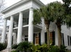 A large white colonial-style house with tall columns, a balcony, and surrounded by lush green palm trees and plants.
