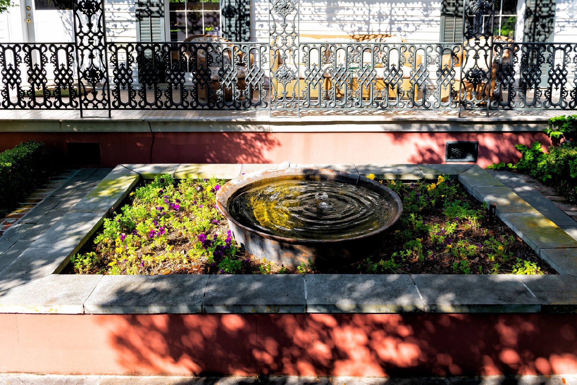 A circular stone fountain with rippling water sits in a small, rectangular garden bed surrounded by green plants and colorful flowers, with a wrought-iron fence and porch in the background.