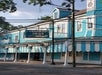 A bright blue building with white and blue striped awnings houses Commander's Palace Restaurant. A blue and white sign with gold text denotes the restaurant's name. Trees and utility lines in the background.