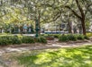 A city park with a tiered fountain in the center, surrounded by trees with hanging moss, and a trolley passing by on the street in the background.