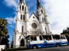 Tour bus in front of a large white gothic-style cathedral with two tall spires on a sunny day.