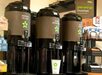 Three coffee dispensers labeled Colombian Rich Roast, Arabian Premium Blend, and Hot Water stand on a counter beside a stack of cups and a sign labeled "grab-and-go breakfast.