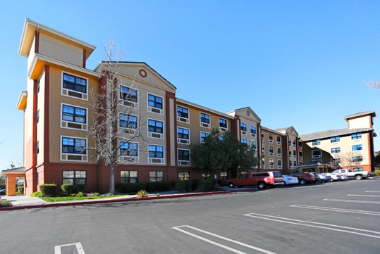 A four-story hotel building with a red and beige exterior, large windows, and a parking lot in front under a clear blue sky.