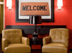 Two yellow armchairs flank a black table with a lamp. Above, a large framed 'WELCOME' doormat hangs on a red wall with two wall lamps.
