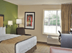 Hotel room with a bed, armchair, desk, and window. Walls are painted green and beige, with framed artwork. Natural light comes through the window with closed curtains.