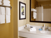 Bathroom with a mirror above a sink, towels on a rack and counter, soap, tissues, and a framed picture on a yellow wall.