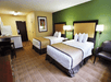 Hotel room with two double beds, a green accent wall, chairs, bedside lamps, a kitchenette, and a table with chairs on a carpeted floor.