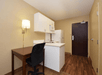 A kitchenette with a fridge, microwave, and sink is next to a dark door. A desk with a chair and a wall-mounted lamp is on the side. The floor is wood, and walls are beige.