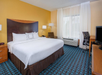 Hotel room with a large bed, nightstands, lamps, a chair, desk with a chair, flat-screen TV, and a window with white curtains. The walls are a mix of yellow and off-white.