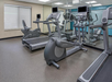 A gym room with exercise equipment, including treadmills, elliptical machines, and a stationary bike, features mirrored walls and a window with a closed blind.