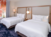 Hotel room with two double beds, white bedding, a nightstand with a lamp and phone, and patterned curtains.