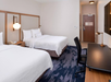 Hotel room with two double beds, a flat-screen TV, desk, chair, and a door with a notice. Room has neutral tones and a blue-patterned carpet.