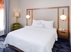 A hotel room with a neatly made double bed, two wall-mounted lamps, a nightstand with a phone, a green chair, and a standing lamp on a carpeted floor.
