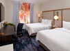 A hotel room with two double beds, a desk with a chair, a TV, and a window with colorful curtains.