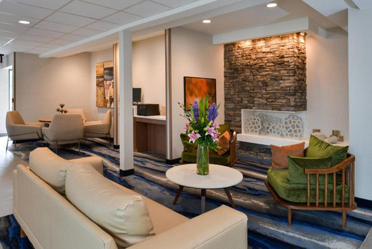 A modern lounge area with beige sofas, stone accent wall, floral arrangement on a table, and abstract artwork. The room features blue carpet and overhead lighting.