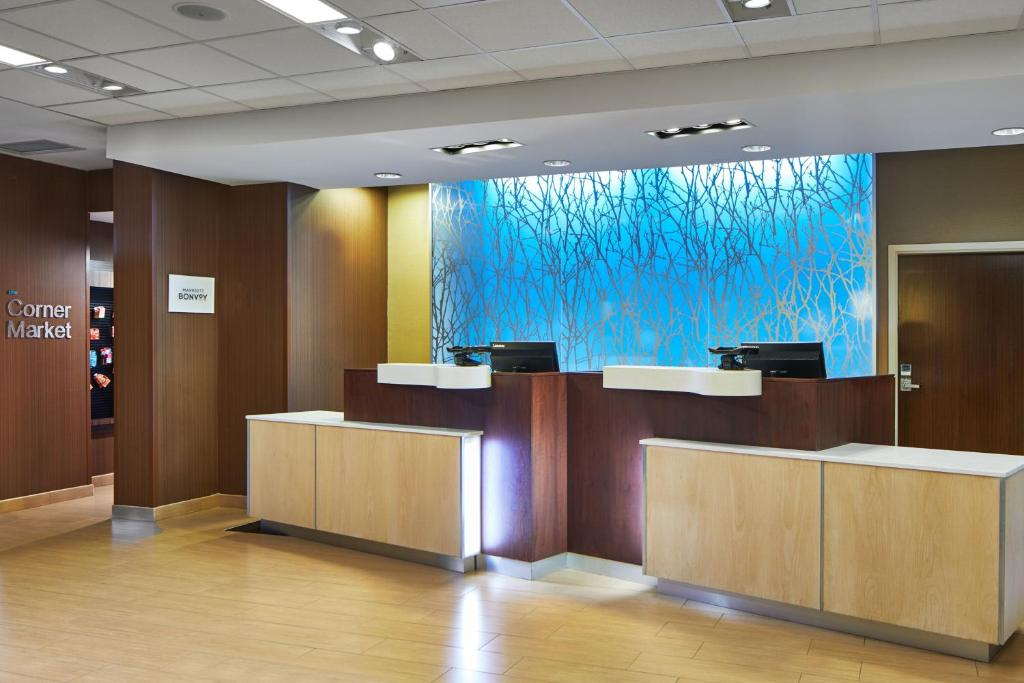 Hotel reception area with a modern design. Two wooden desks with computers are in front of a blue and white decorative wall. A sign for "Corner Market" is visible to the left.