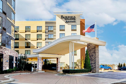 Fairfield Inn & Suites by Marriott exterior with a modern entrance, an American flag flying above, and a blue car parked nearby under a partly cloudy sky.