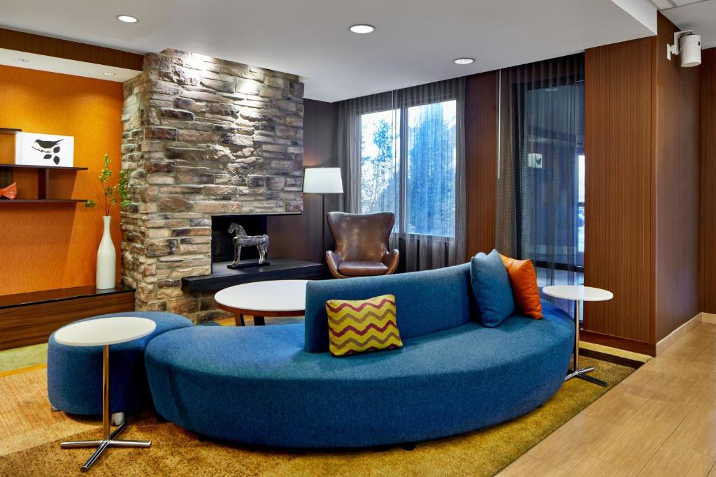 A modern hotel lobby with a blue curved sofa, pillows, and round tables. A stone fireplace and leather chair are in the background, with orange accents around the room.
