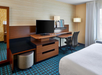 Hotel room with a double bed, desk, chair, TV, floor lamp, and window with curtains. Walls have a striped pattern, and the carpet is blue with a geometric design.