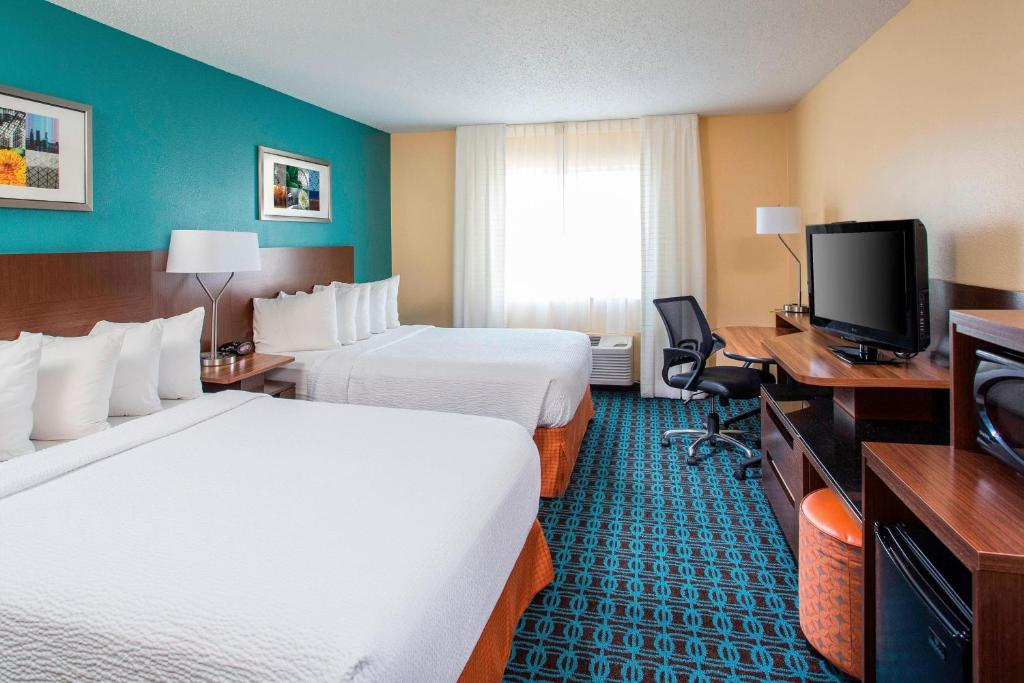Hotel room with two double beds, a desk with chair, flat-screen TV, mini fridge, microwave, and window with sheer curtains; walls painted teal and beige with framed artwork.
