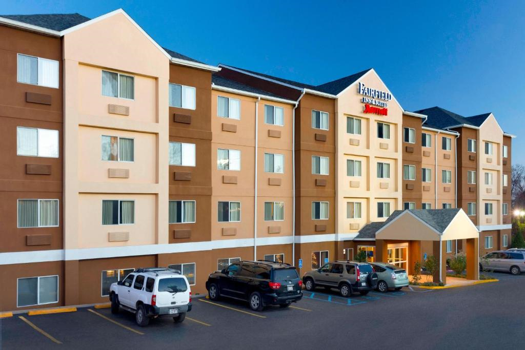 Four-story hotel building with tan and brown exterior, multiple windows, and a covered entrance; several cars are parked in front in a lot.