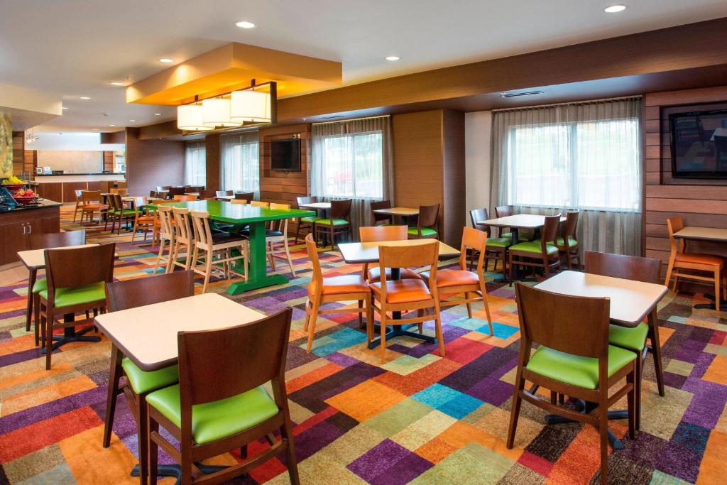 A brightly lit dining area with wooden tables and chairs, colorful patterned carpet, large windows, and modern ceiling lights.