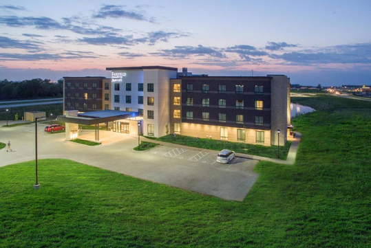 A modern four-story hotel building with a lit entrance and parking lot, surrounded by open grassy fields at dusk.