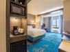Hotel room with a bed, desk, and window. In the foreground, there is a microwave, coffee maker, and mini-fridge on a shelf. The room features blue carpeting and neutral walls.