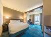 Hotel room with a large bed, bedside tables with lamps, a desk with a chair, and a window with curtains, featuring blue carpet and neutral wall colors.