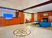 Hotel lobby with brown wood paneling, a reception desk, seating area with orange chairs and a blue sofa, and a tile floor with a decorative circular pattern.