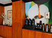 Coffee station with three dispensers and various cups, set against a large coffee-themed mural, next to a vase with flowers and a water container on wooden cabinets.
