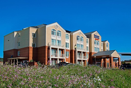 A four-story hotel with a red and beige exterior stands behind a grassy field with small pink and white flowers under a clear blue sky.