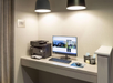 A small office nook with a printer, a computer monitor displaying a hotel website, a keyboard, a mouse, and supplies under two hanging lights.