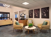 Modern waiting area with two beige chairs, a round white table, and wall art. Coffee station visible in the background. Neutral tones and minimalist decor.