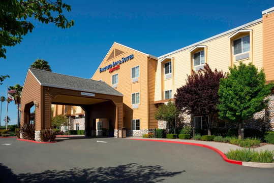 A beige hotel exterior with a covered entrance labeled "ENTRANCE" and a "FAIRFIELD INN & SUITES" sign. The building has three floors, surrounded by trees and landscaping.