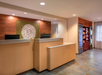 Hotel reception area with wooden front desk, a decorative wall piece behind, and a snack bar with refrigerators on the right.