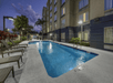 An outdoor swimming pool with the Fairfield Inn & Suites on the right and lounge chairs with bushes behind them on the left at night.