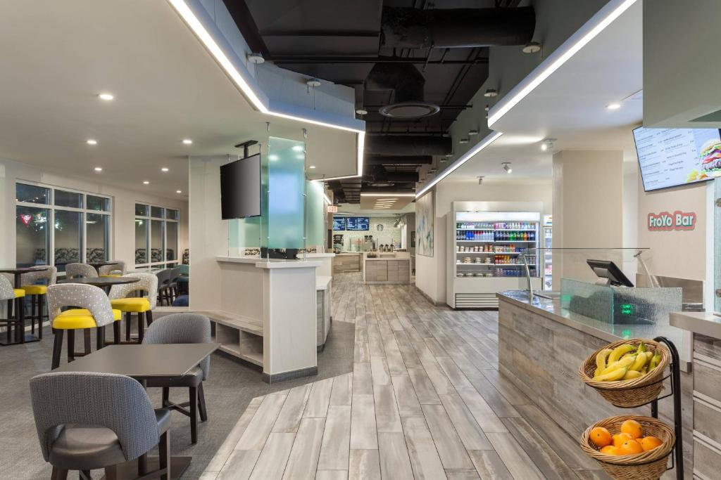 A modern café interior with gray chairs, yellow stools, a counter, digital menu boards, refrigerated display, and baskets of fruit on the right.