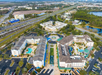 Aerial view of a hotel complex with pools, courtyards, parking lots, and surrounding roads, set near a highway and green spaces.