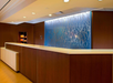 Modern hotel reception desk with a wood finish, white countertop, and a decorative blue glass panel in the background. A small snack area is visible to the left.